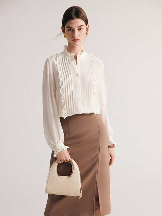 A woman wearing a white pleated blouse with ruffle trim, long sleeves, and a stand collar, paired with a brown skirt. She is holding a beige handbag.