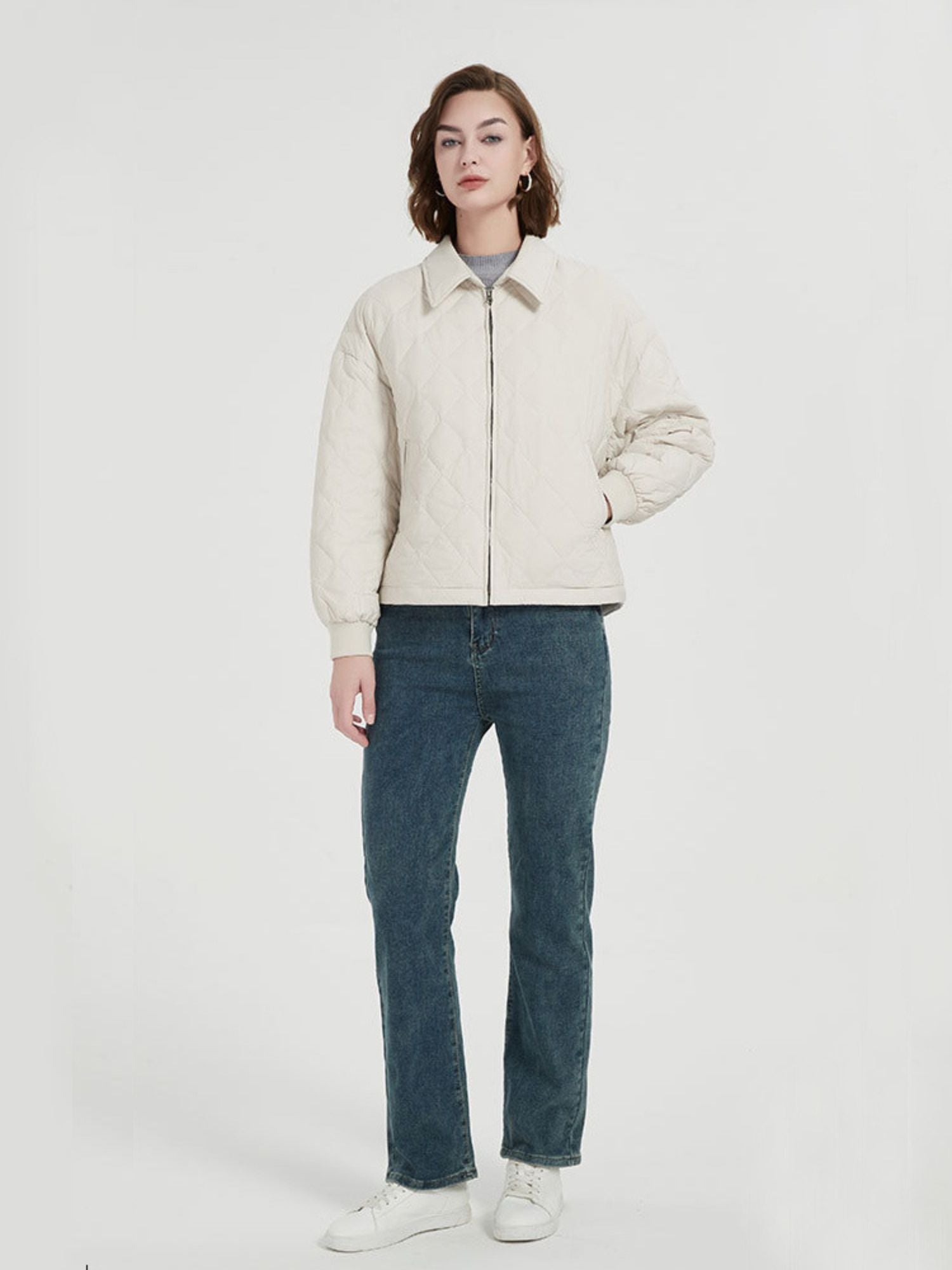 A female model wearing a beige thick quilted down coat with pockets, paired with a grey top and blue jeans.