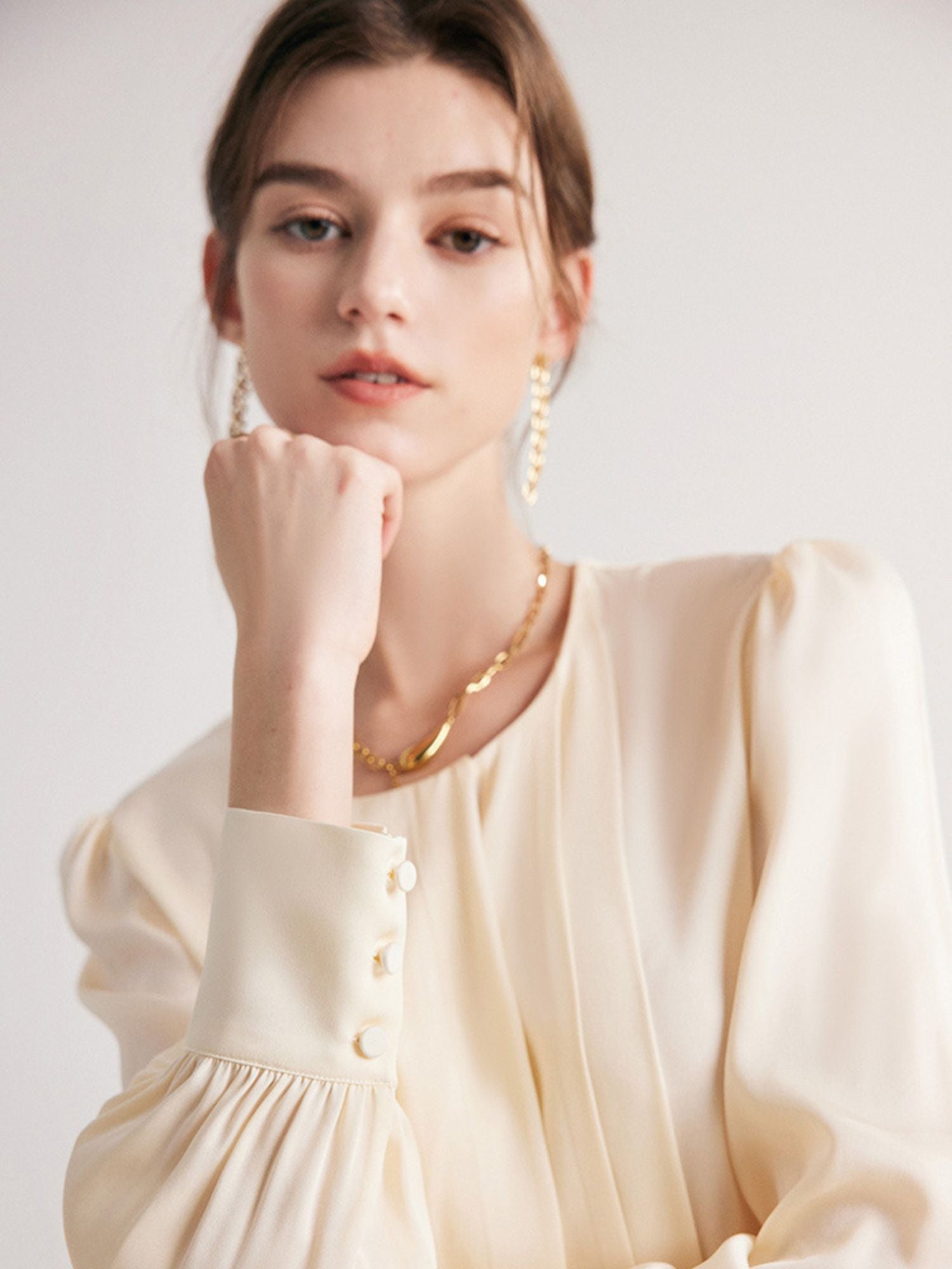 A woman wearing a round neck silk blouse with pleated details, long sleeves, and buttoned cuffs in a cream color.