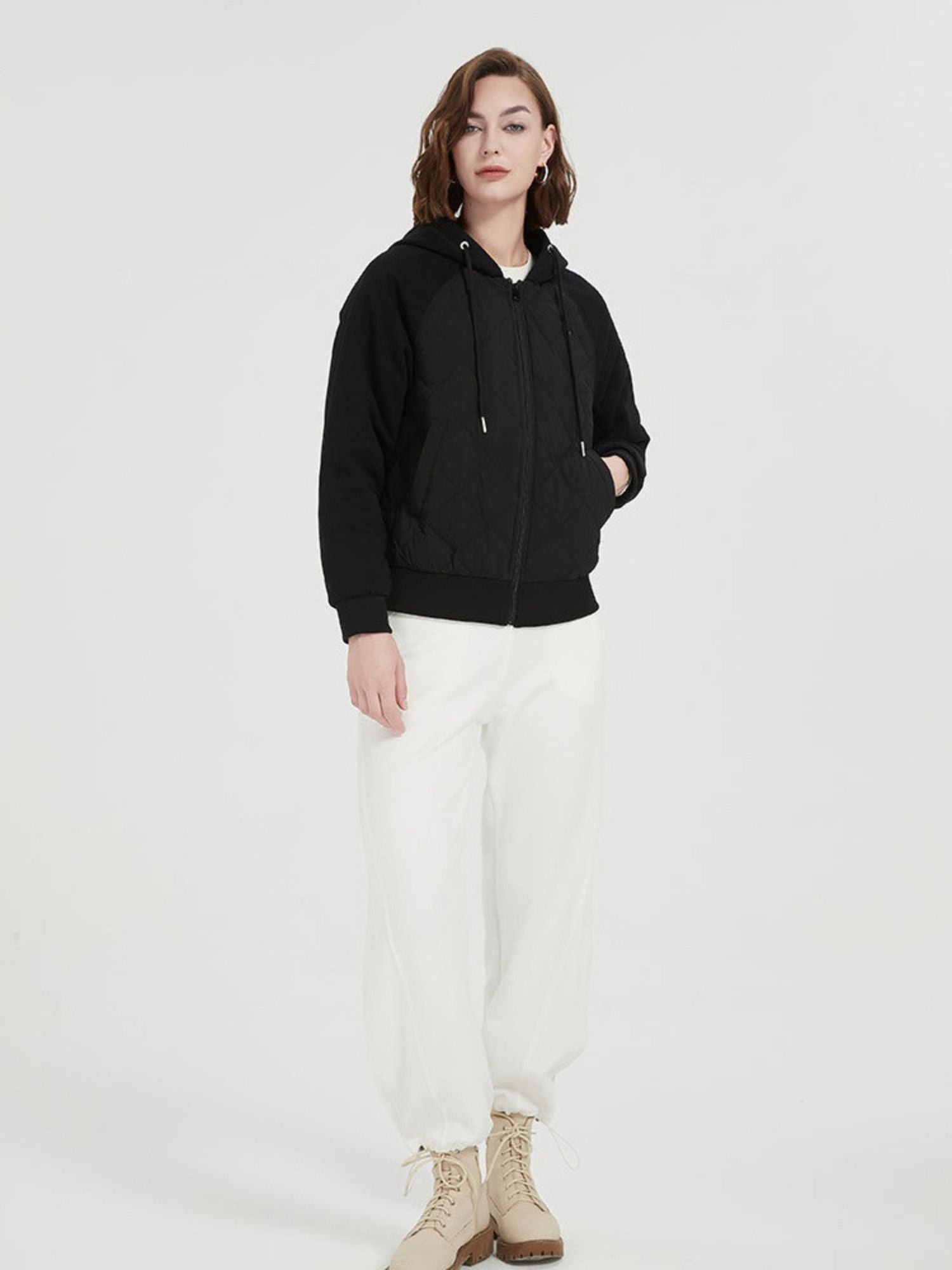 A female model wearing a black quilted down jacket with a hood, paired with a white crop top and white pants.
