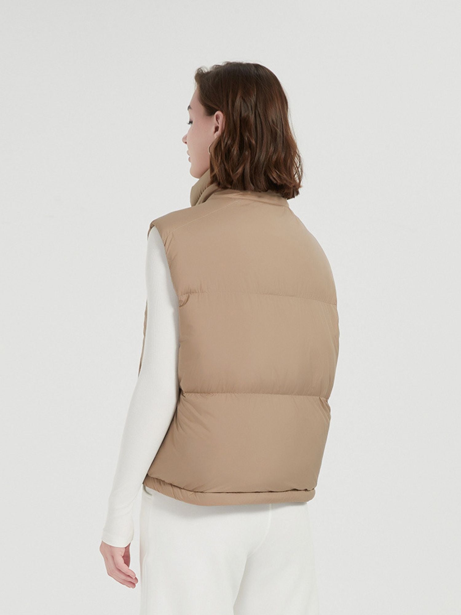 A female model wearing a beige quilted down vest with a stand collar, sleeveless design, and a full zip.