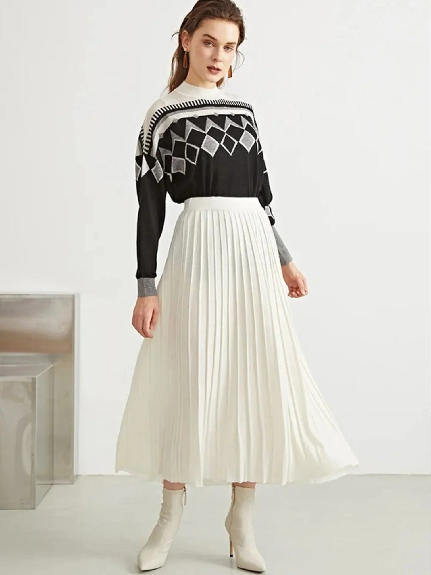 A woman wearing a black and white geometric patterned sweater paired with a pleated long skirt in a beige wool blend.