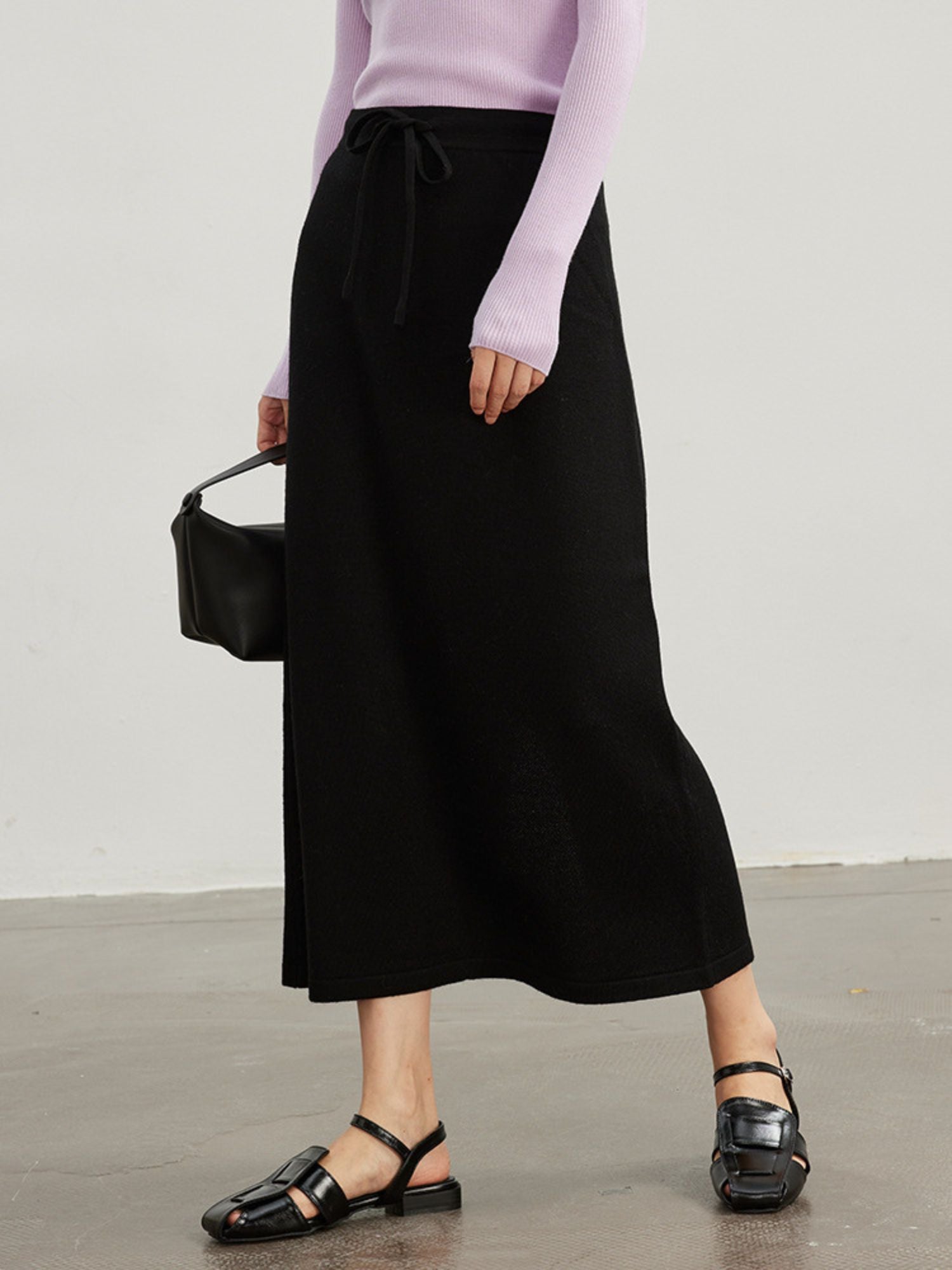 A woman wearing a midi-length black skirt with an adjustable drawstring waist, paired with a purple turtleneck top and black sandals.