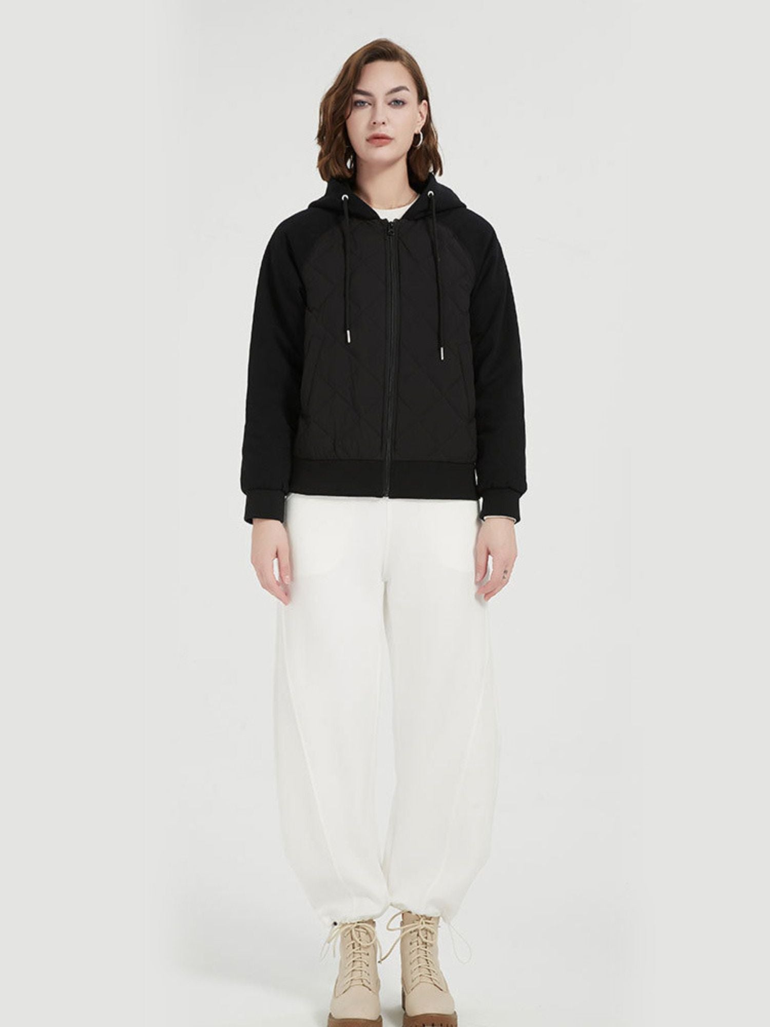 A female model wearing a black quilted down jacket with a hood, paired with a white crop top and white pants.
