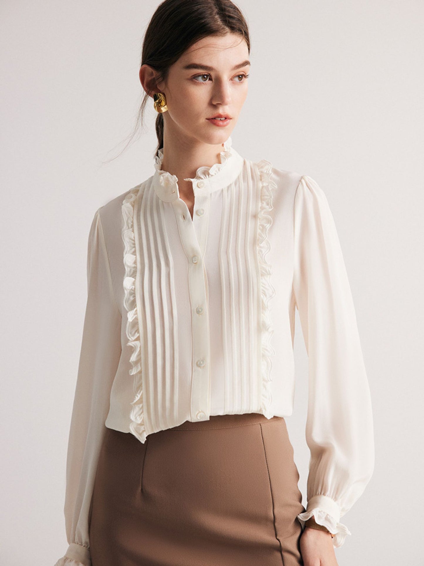 A woman wearing a white pleated blouse with ruffle trim, long sleeves, and a stand collar, paired with a brown skirt. She is holding a beige handbag.