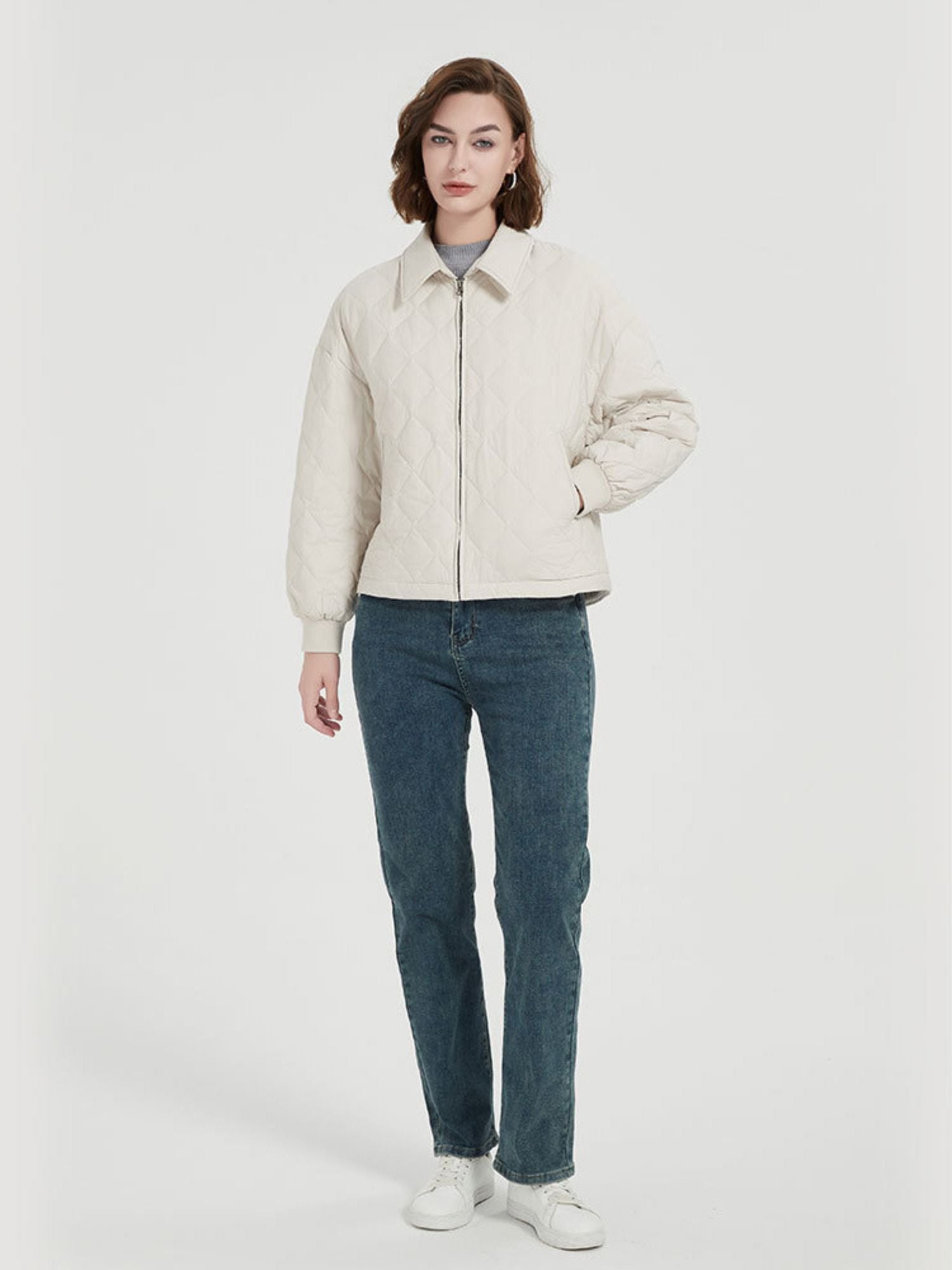 A female model wearing a beige thick quilted down coat with pockets, paired with a grey top and blue jeans.