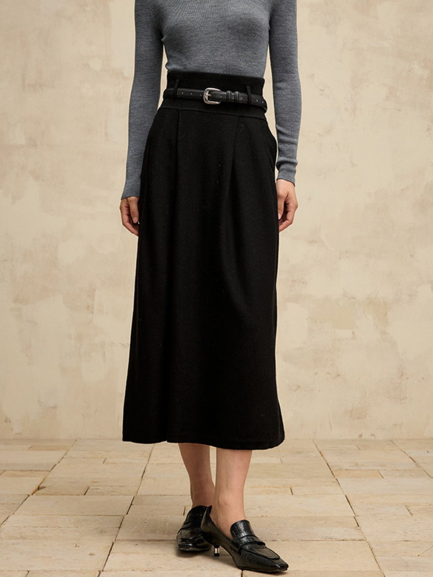 A woman wearing a high-waisted midi length black skirt with a flared silhouette and a belt detail. She has paired it with a gray turtleneck sweater.