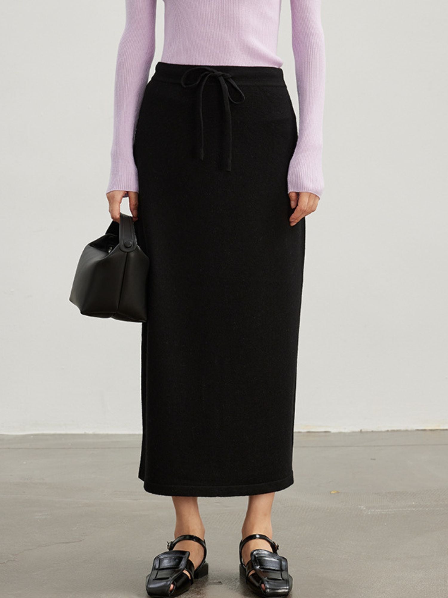 A woman wearing a midi-length black skirt with an adjustable drawstring waist, paired with a purple turtleneck top and black sandals.