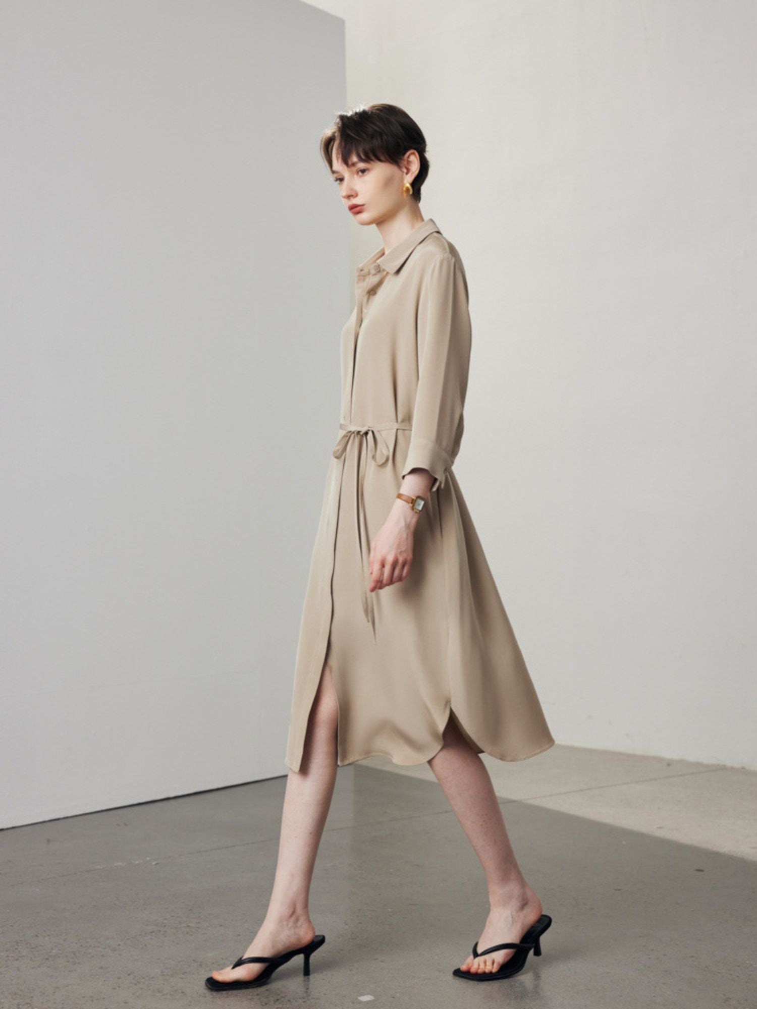 A woman with short hair wearing a lose dress shirt in khaki with black sandals.