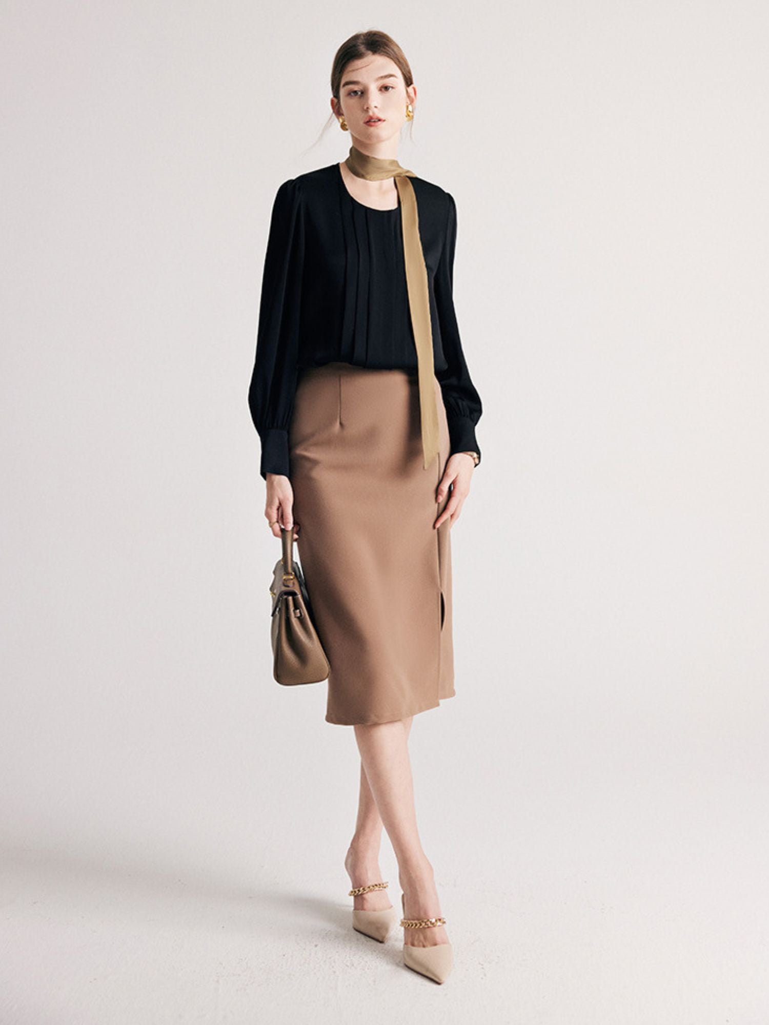 A woman wearing a round neck silk blouse with pleated details, long sleeves, and buttoned cuffs with a brown skirt.
