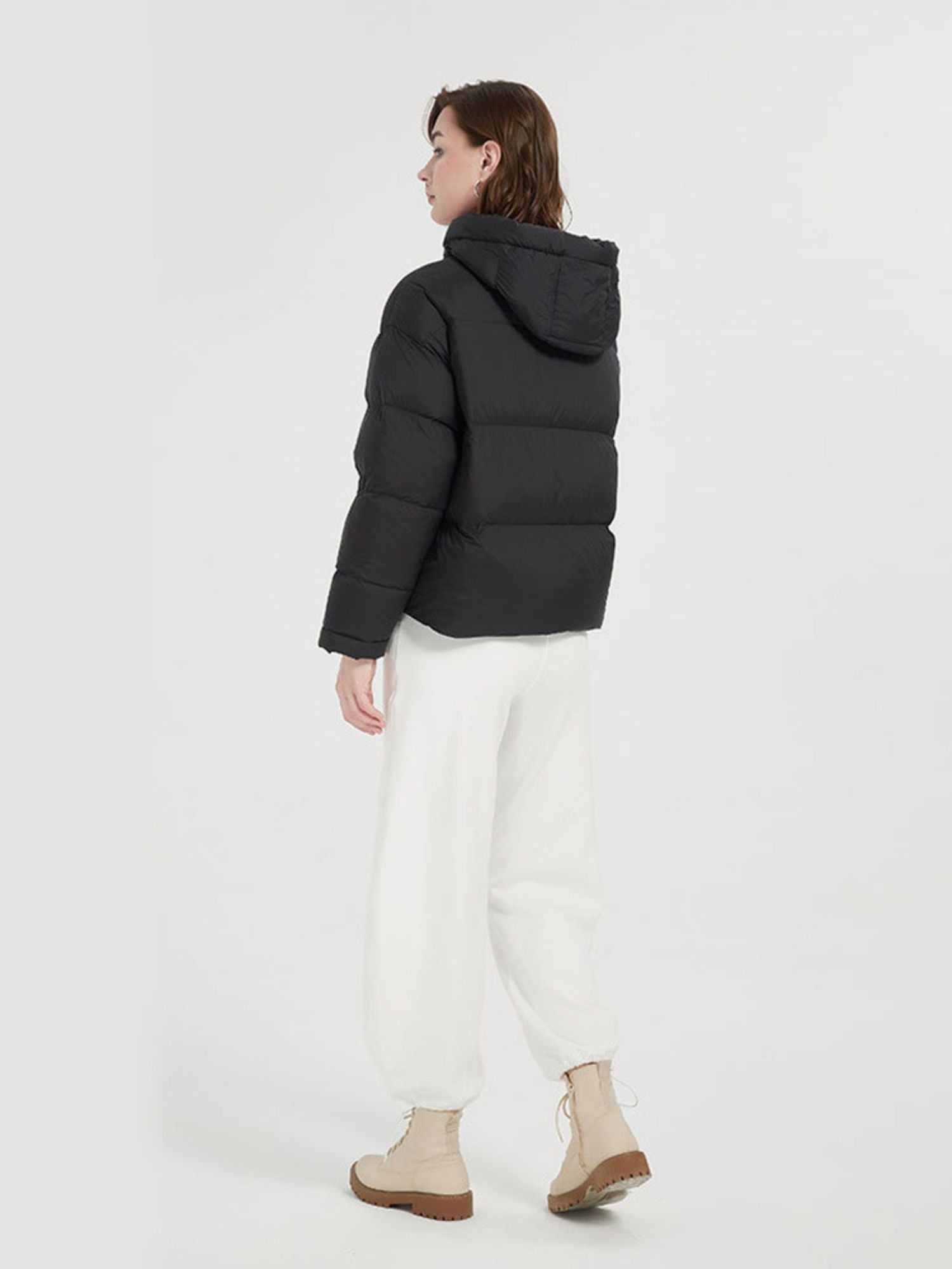 A female model wearing a black quilted down jacket with a hood and pockets, paired with white pants and beige boots.